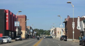 Street with cars, motorcycles, and pedestrians in a small town lined with buildings and storefronts, including MVP sign and flags.